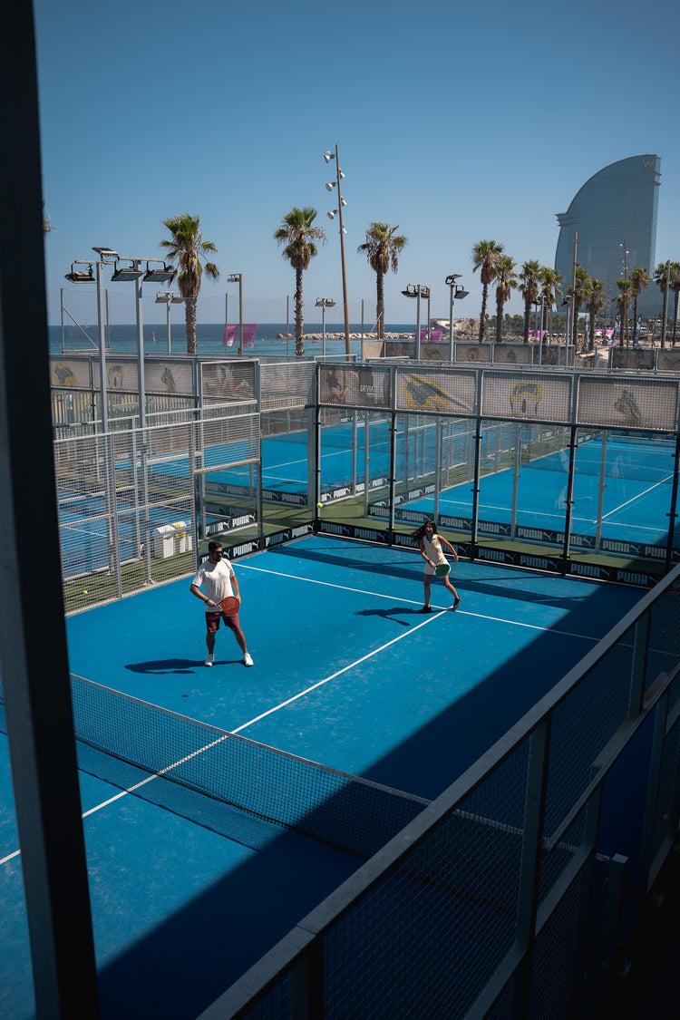 Two players on a blue court by the sea, seen from above